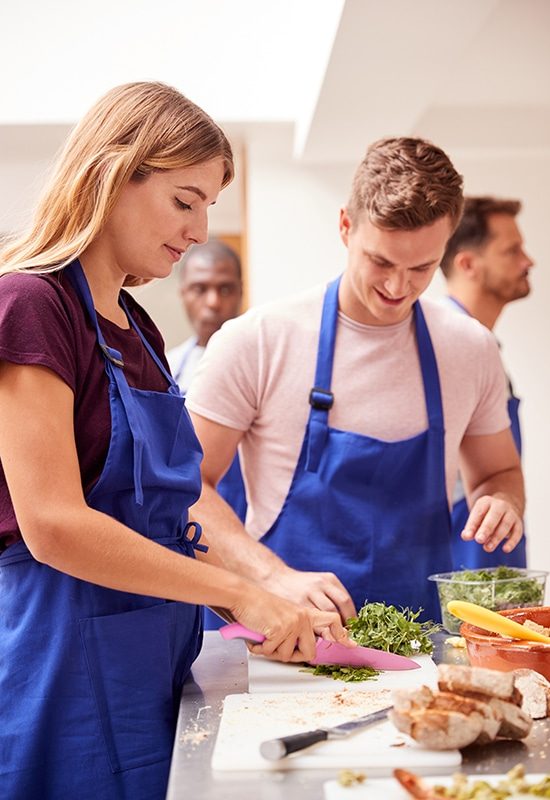 Male And Female Adult Students Preparing Ingredients For Dish In Kitchen Cookery Class